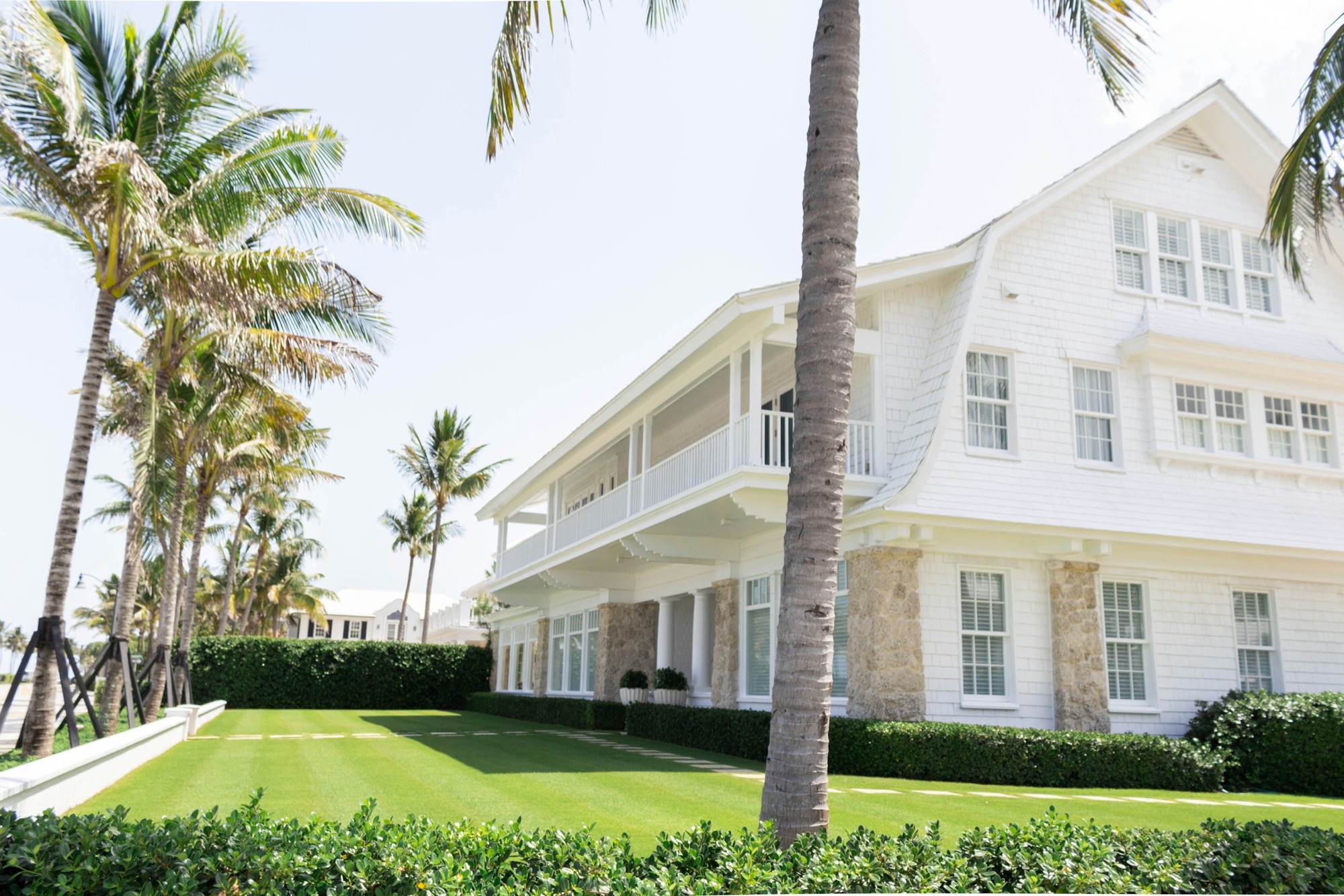 White shingle style house behind clipped hedges and palm trees