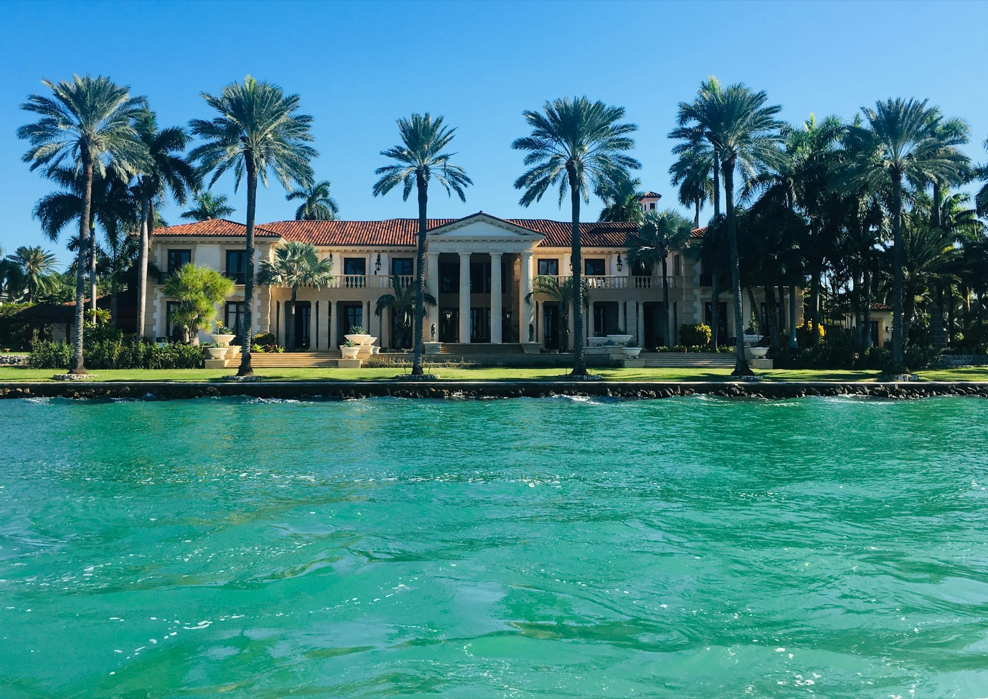 Mediterranean waterfront house with a palm allee seen across turquoise water