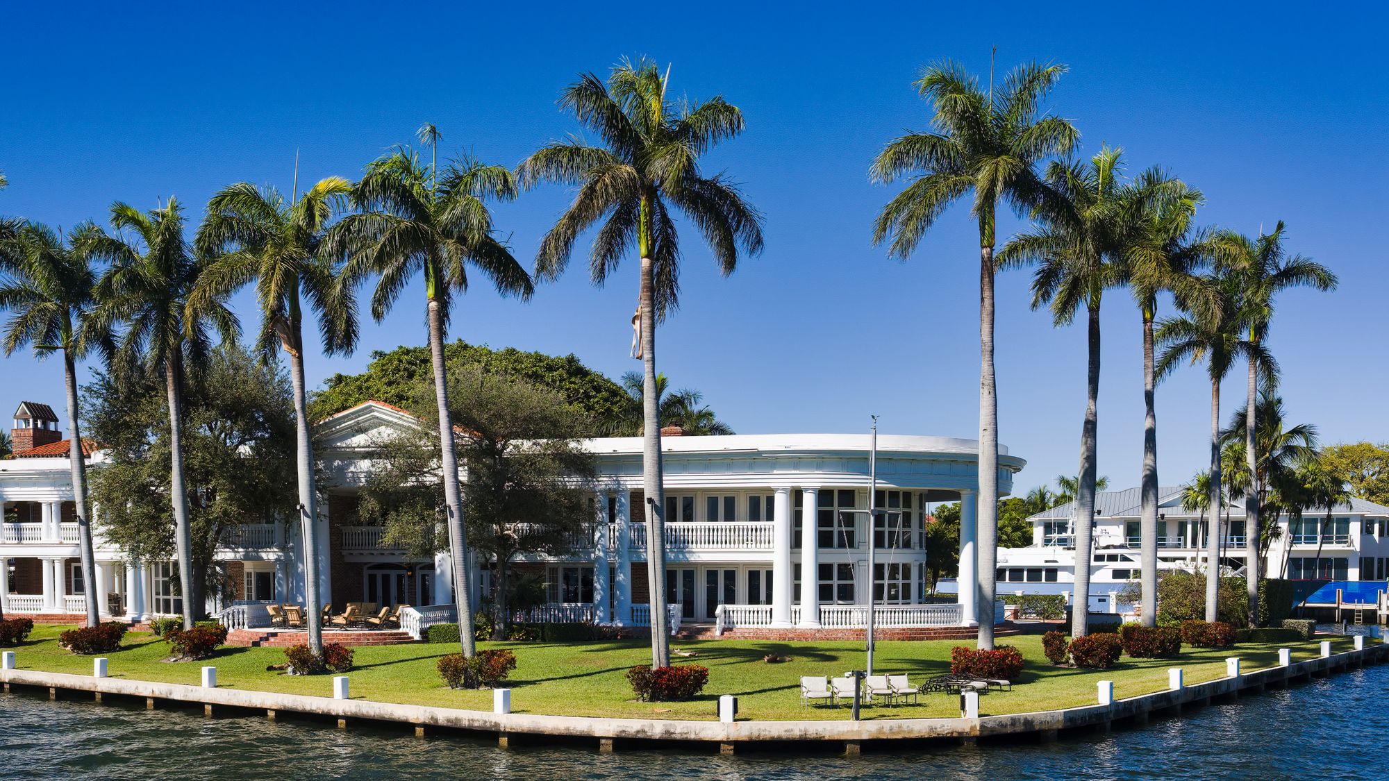 White waterfront estate with balconies, a lawn and tall palms beside a canal